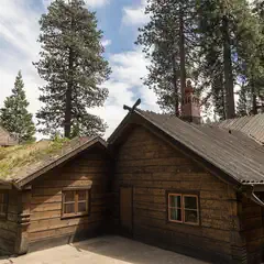 A rustic wooden cabin with a grass-covered roof in front of a forested area with towering evergreen trees under a partly cloudy sky.