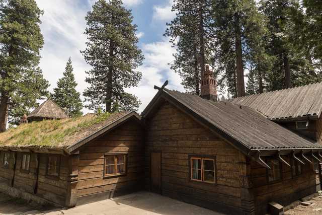A rustic wooden cabin with a grass-covered roof in front of a forested area with towering evergreen trees under a partly cloudy sky.