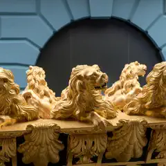 Five gilded lion sculptures rest on an ornate golden railing, set against a blue paneled wall with a dark arched opening.