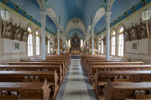A church interior with high ceiling decorated in blue and gold, wooden pews, and stained glass windows providing natural light.