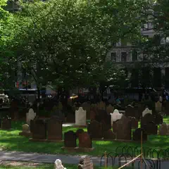 A cemetery filled with weathered headstones of varying shapes and sizes, surrounded by trees and urban buildings in the background.