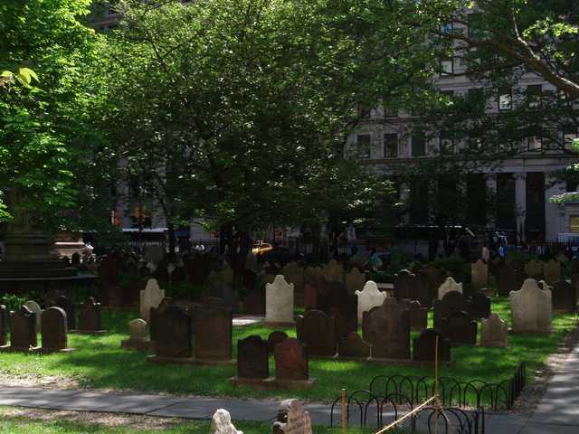 A cemetery filled with weathered headstones of varying shapes and sizes, surrounded by trees and urban buildings in the background.