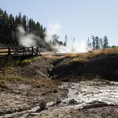 A wooden bridge over a rocky stream, with steam rising from a geothermal vent nearby and trees on the hillside in the background.