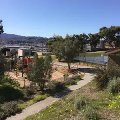 An elevated park overlooks a city, featuring trees, grassy areas, and a playground with red equipment.