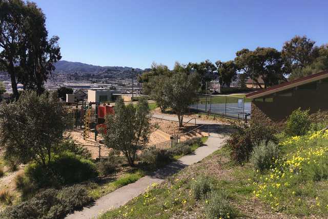 An elevated park overlooks a city, featuring trees, grassy areas, and a playground with red equipment.