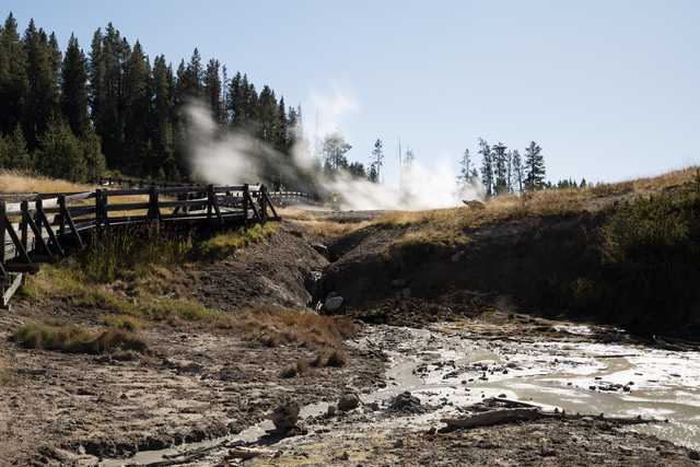 A wooden bridge over a rocky stream, with steam rising from a geothermal vent nearby and trees on the hillside in the background.