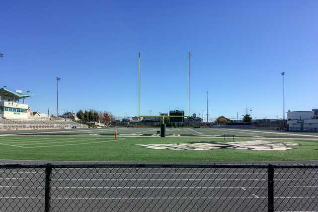 A football field with goalposts and a scoreboard, viewed from behind a chain-link fence.