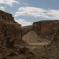 A dirt road curves through a narrow canyon with steep, reddish-brown rock walls and a blue sky overhead.