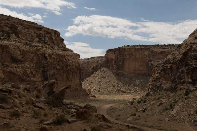 A dirt road curves through a narrow canyon with steep, reddish-brown rock walls and a blue sky overhead.