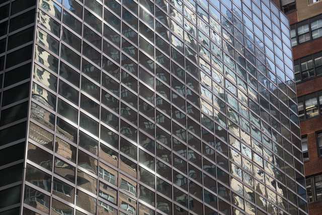 A modern glass facade reflects the distorted image of a nearby brick building.