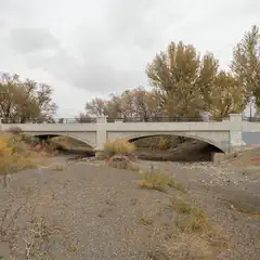 A concrete bridge with a metal railing crossing over a dry riverbed with sparse vegetation and rocky terrain.