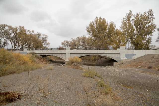 A concrete bridge with a metal railing crossing over a dry riverbed with sparse vegetation and rocky terrain.