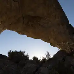 A natural rock arch frames a clear sky above rugged terrain and scattered vegetation.