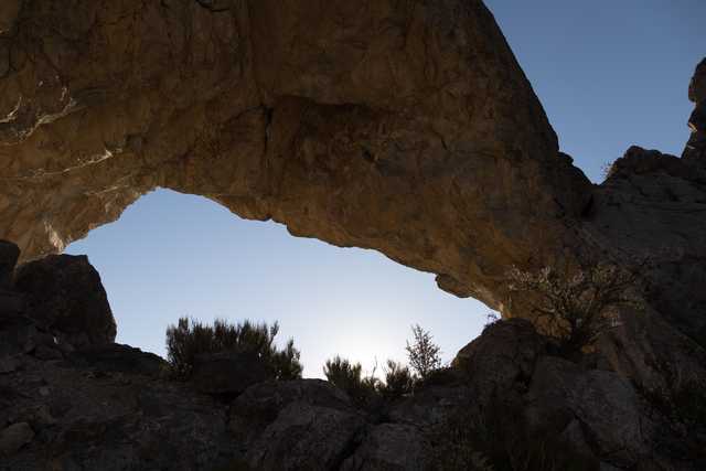 A natural rock arch frames a clear sky above rugged terrain and scattered vegetation.