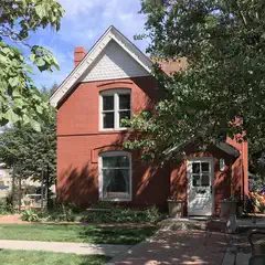 A red two-story house with a white door and window frames, surrounded by grass and trees, under a partly cloudy sky.