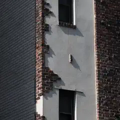Two vertically aligned windows are set into a light-colored stucco wall, bordered by dark brickwork with irregular, stepped masonry.