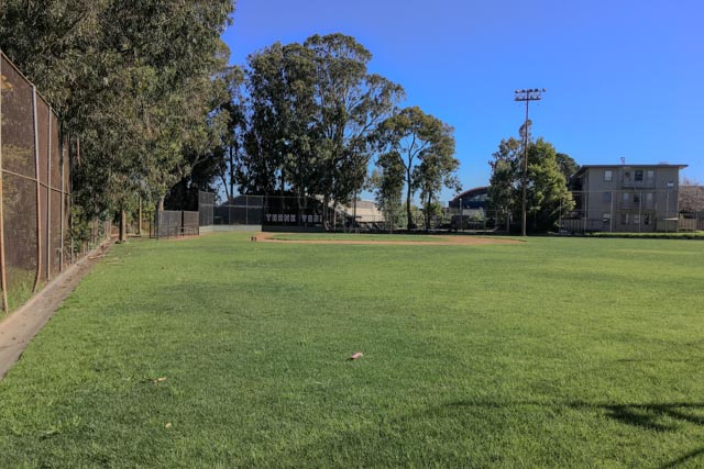 A baseball field occupies a large grassy area with a backstop, trees, and a building visible in the distance.