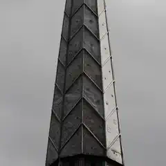 A tall, pointed spire constructed of weathered, diamond-patterned panels rises against a grey sky.