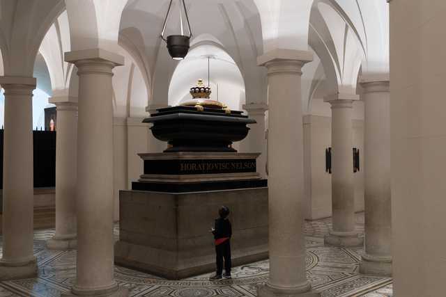 A child stands before the tomb of Horatio Viscount Nelson in a vaulted hall with columns and a patterned floor.