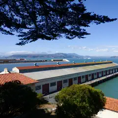 A long industrial building with red doors sits along a waterfront, with a large bridge and distant hills visible across the water under a clear blue sky.