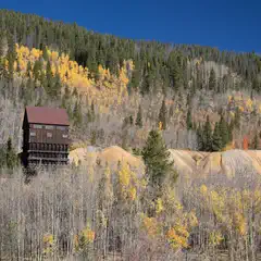 A wooden structure with a red roof is nestled in the mountains. It's surrounded by trees with yellow leaves indicating autumn. The sky above is clear and blue.