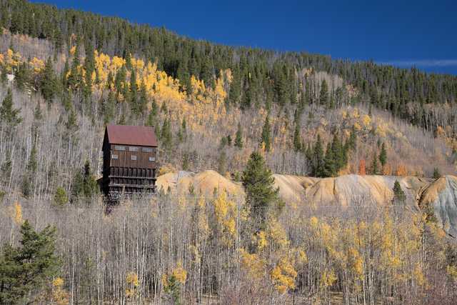 A wooden structure with a red roof is nestled in the mountains. It's surrounded by trees with yellow leaves indicating autumn. The sky above is clear and blue.