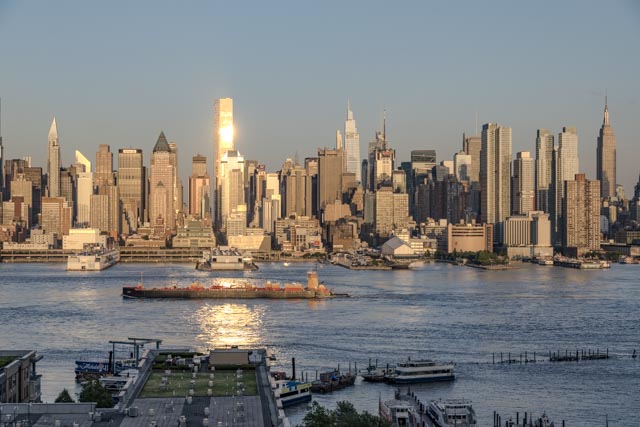 The sun reflects off a skyscraper in Manhattan's skyline, with a barge and several boats visible on the water in the foreground.