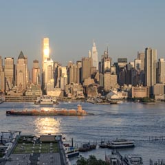 The sun reflects off a skyscraper in Manhattan's skyline, with a barge and several boats visible on the water in the foreground.