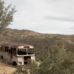 An old, rusted bus parked on a dirt road surrounded by desert scrub brush and mountains in the background.