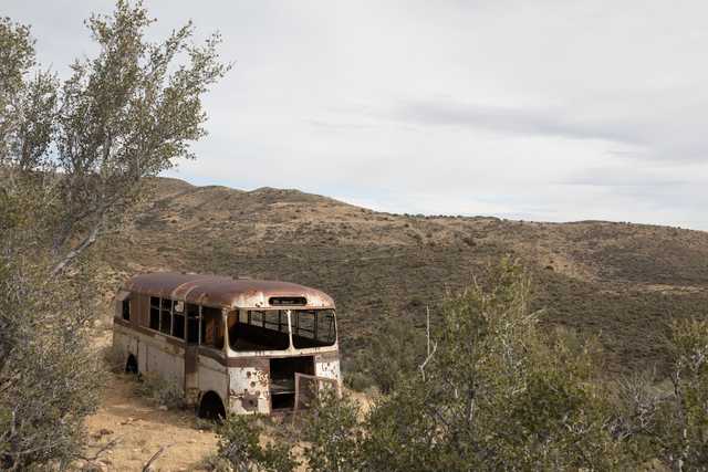 An old, rusted bus parked on a dirt road surrounded by desert scrub brush and mountains in the background.