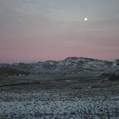 A snow-covered landscape with mountains in the distance under a pink and blue sky. The full moon is visible in the sky.