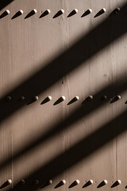 A wooden door with metal studs and bolts, partially shaded by sunlight filtering through slats above it.