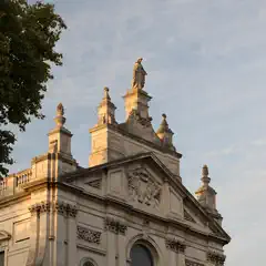A neoclassical building features ornate stonework, decorative urns, and a central statue atop its roofline.