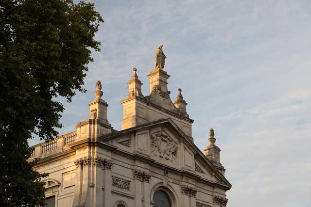 A neoclassical building features ornate stonework, decorative urns, and a central statue atop its roofline.