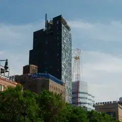 A tall building under construction, surrounded by other buildings and cranes, with trees visible in the foreground.