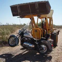 A yellow backhoe is lifting a gray motorcycle off the ground on a dirt road in a desert landscape.