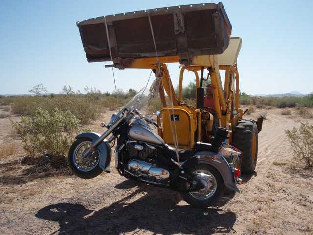 A yellow backhoe is lifting a gray motorcycle off the ground on a dirt road in a desert landscape.