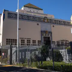 A beige building with decorative tilework and a Court of Honor sign is enclosed by a black metal fence, with a circular sign on a pole to the right.