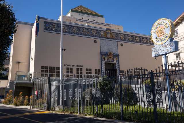 A beige building with decorative tilework and a Court of Honor sign is enclosed by a black metal fence, with a circular sign on a pole to the right.