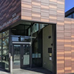 An entrance to a modern library building, featuring large windows and a textured brown facade with the name of the institution displayed above the doors.