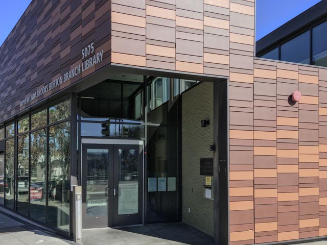 An entrance to a modern library building, featuring large windows and a textured brown facade with the name of the institution displayed above the doors.