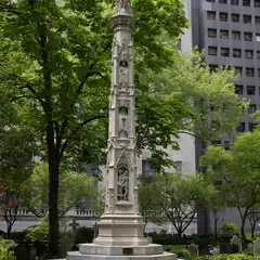A tall, ornate stone cross stands prominently in a well-maintained garden, surrounded by trees and grave markers, with modern buildings in the background.