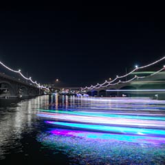 A waterway is spanned by two bridges and lined with strings of lights; illuminated boats create streaks of purple and white on the dark water.
