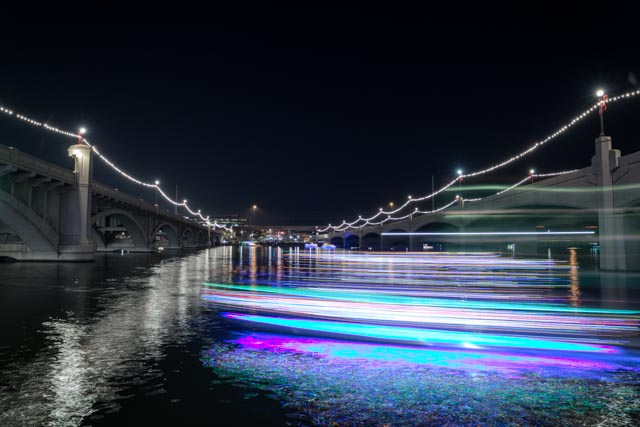 A waterway is spanned by two bridges and lined with strings of lights; illuminated boats create streaks of purple and white on the dark water.