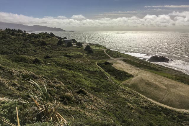 A coastal landscape under a partly cloudy sky, featuring grassy hillsides leading to a sandy beach with waves lapping at its edge and a rocky coastline beyond.