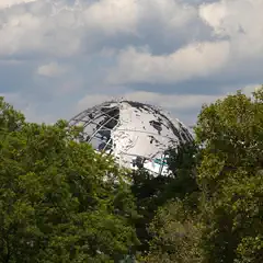 A large, metallic globe with a skeletal framework and fragmented surface rises above a dense canopy of green trees under a cloudy sky.