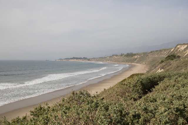 A coastal landscape features a sandy beach, gentle waves, and a bluff covered in vegetation under a hazy sky.