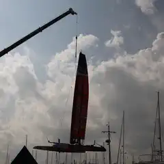 A multihull boat with a tall red sail hangs from a crane above a marina filled with sailboats.