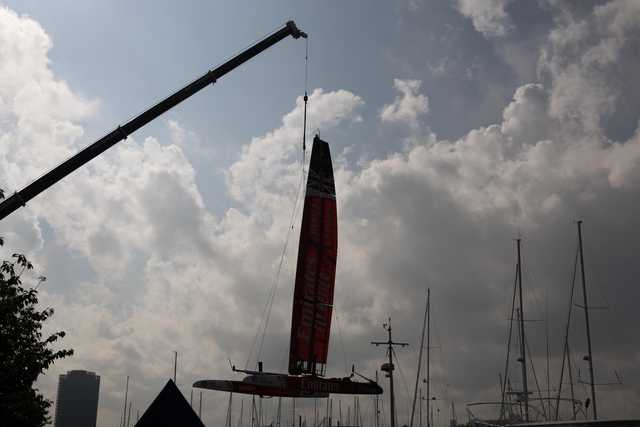 A multihull boat with a tall red sail hangs from a crane above a marina filled with sailboats.