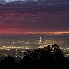 A panoramic nighttime view of a cityscape with illuminated buildings, a bridge, and a vibrant red and purple sky.